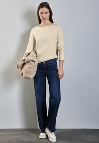 Crèmekleurig longsleeve shirt, donkerblauwe rechte jeans en witte sneakers. Model houdt een beige fluffy jas vast, tegen een lichtgrijze achtergrond.