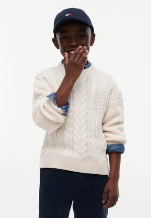 Dreng iført en mørk kasket, cremefarvet kabelstrikket sweater over en denimskjorte, smilende med den ene hånd delvist dækkende sin mund.