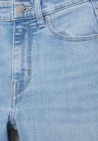 Primo piano di jeans in denim azzurro chiaro che mostra la tasca frontale, il passante per la cintura, il bottone di metallo e i dettagli delle cuciture a contrasto.