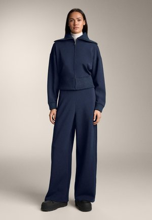 Mornarsko plavi trenirka s kratkom jaknom na patentni zatvarač i širokim hlačama. Rebrasti manšeti i rub. Glatki, mekani materijal s opuštenim krojem.