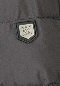 Sort stofmærke med et sølvfarvet skjolddesign, der viser "NMT 97" og krydsede linjer. Glat tekstur med en broderet kant.