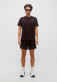 Νέος άνδρας που φοράει μαύρα αθλητικά ρούχα, γυαλιά ηλίου, λευκές κάλτσες και λευκά αθλητικά παπούτσια, στέκεται μπροστά σε απλό φόντο.