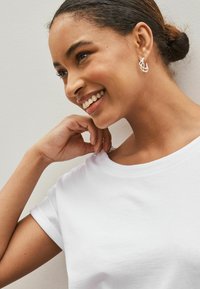 Lachende vrouw met haar in een knot draagt zilveren oorringen en een wit T-shirt, waarbij ze haar hand bij haar nek houdt tegen een neutrale achtergrond.