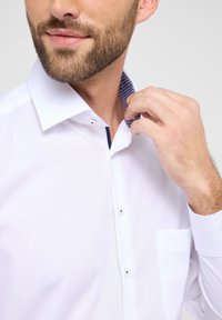 Weißes Hemd mit Knopfleiste, das einen kontrastierenden navy-blauen und karierten Innenkragen aufweist. Glatte Textur, mit einer Brusttasche und sichtbaren Knöpfen.