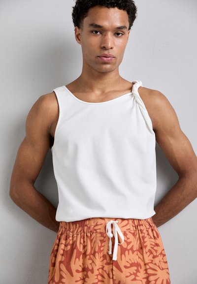 White sleeveless top with a twisted detail on one shoulder, paired with orange shorts featuring a leaf pattern and elastic waistband with drawstring.