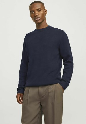 Maglione girocollo navy in maglia testurizzata, con maniche lunghe e polsini a coste, abbinato a pantaloni plissettati color marrone chiaro.