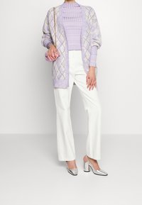 Lila-musterter Cardigan mit Rautendesign über einem gerippten lila Top, kombiniert mit weißen weit geschnittenen Hosen und silbernen Blockabsatzschuhen.