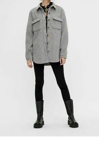 Graue Wollhemdjacke mit zwei Brusttaschen, fünf Knöpfen und abgerundetem Saum. Kombiniert mit schwarzen Leggings und klobigen schwarzen Stiefeln.