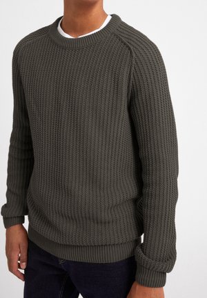 Uomo che indossa un maglione di maglia strutturata verde oliva scuro sopra una canottiera bianca e jeans scuri, in piedi davanti a uno sfondo bianco semplice.