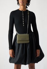 Grønn skinn crossbody bag med glossy finish, kjede-reim og preget "REPLAY"-logo. Modellen har på seg en ribbet svart topp og ett svart skjørt.