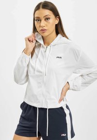 Valkoinen kevyt vetoketjullinen takki hupulla ja joustavilla hihansuilla, jossa on Fila-logo. Yhdistetty tummansinisiin shortsseihin, joissa on valkoiset sivuviivat.