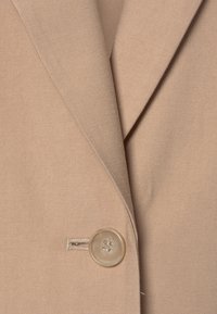 Beżowa marynarka z teksturowanej tkaniny, z wyciętym kołnierzem i wyróżniającym się okrągłym guzikiem z czterema dziurkami blisko otworu.