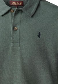 Polo vert texturé avec deux boutons marron, col plié et petit logo noir brodé sur le côté gauche de la poitrine.