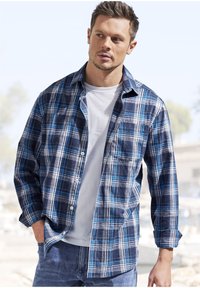 Plaid shirt features navy, white, and light blue colors, button closure, rolled sleeves, and a chest pocket. Worn over a white t-shirt.