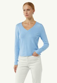 Hellblaues, langärmliges V-Ausschnitt-Pullover aus weichem Material. Mit gerippten Bündchen und Saum. Getragen mit weißen Hosen, Hände in den Taschen.