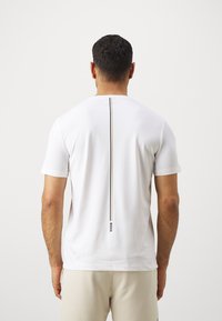 Valge lühikeste varrukatega T-särk, mille taga on sirge must ja kuldne vertikaalne triipude muster ning logo detail hemmi lähedal. Sile tekstuur.