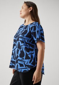 Blue and black patterned short-sleeve top with a crew neck and a slightly longer back hem. Textured fabric with abstract shapes.