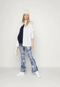 Mulher grávida a usar um gorro branco, uma camisa branca, um top azul marinho, calças azul tie-dye, sapatilhas brancas, segurando uma mala bege, a posar contra um fundo neutro.