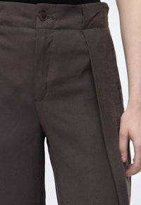 Gros plan sur un pantalon marron foncé avec un bouton et une fermeture éclair à l'avant, porté par une personne dont la main droite repose à côté de la jambe.