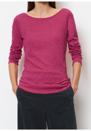 Vrouw met een lange mouw, vervaagde magentakleurige top met een brede halslijn en zwart gestreepte broek, handen in de zakken, tegen een effen achtergrond.