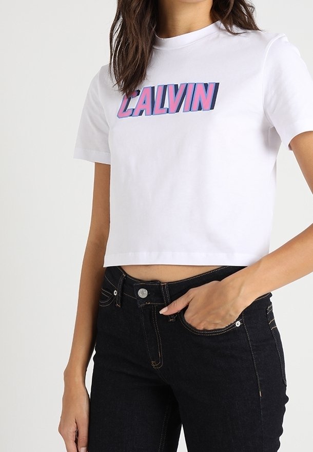 Žena v bílém krátkém tričku s nápisem „CALVIN“ růžovými a modrými písmeny, doplněném o černé džíny s vysokým pasem.