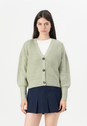 Lysegrønn strikket cardigan med V-hals, fem knapper og ribbestrikkede mansjetter. Kombinert med en marineblå plissert skjørt. Myk tekstur.