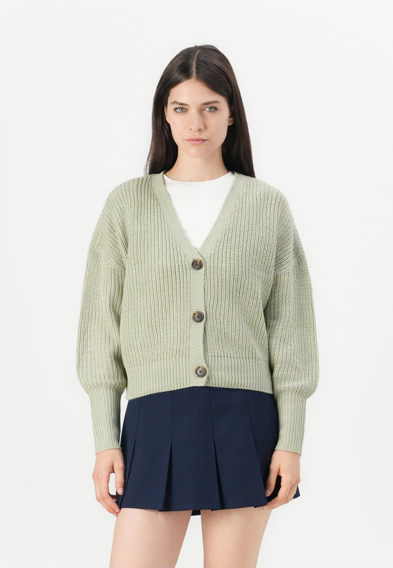 Cardigan tricoté vert clair avec un col en V, cinq boutons et des poignets côtelés. Assorti avec une jupe plissée bleu marine. Texture douce.