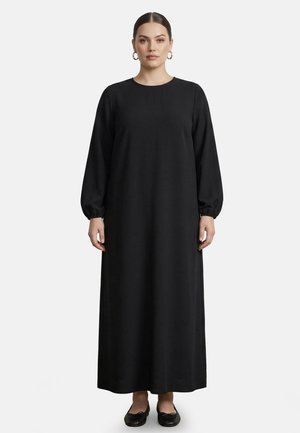 Vrouw staat, draagt een lange zwarte jurk met lange mouwen en zwarte platte schoenen, haar naar achteren getrokken en gouden ringoorbellen.