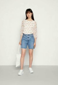 Blusa branca transparente com padrão floral e mangas 3/4, combinada com calções de ganga azuis de corte descontraído e bainha desfiada. Ténis brancos e mala.