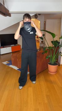 Črna majica, ohlapne črne hlače in črna bandana z belim vzorcem pajkove mreže. V sobi je rastlina v lončku in lesena tla.