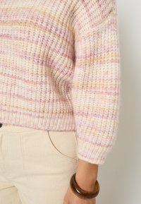 Maglione lavorato a maglia in tonalità di rosa pallido e beige, con una texture a coste e un design corto. Abbinato a pantaloni di colore chiaro e un bracciale rotondo.