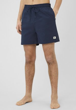 Homme portant un short bleu marine avec taille élastique, cordon de serrage et poches latérales, debout pieds nus devant un fond uni.