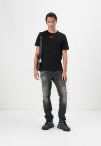 Čierne tričko s krátkym rukávom a červeným logom, spárované so sivými džínsami. Model má na sebe čiernu crossbody tašku a tmavé športové topánky.