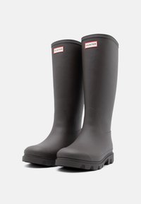 Čierne gumové zatepérové čižmy s hladkým povrchom, okrúhlou špičkou, textúrovanou podrážkou a značkovou etikou v červenej a bielej farbe na vrchnej časti.