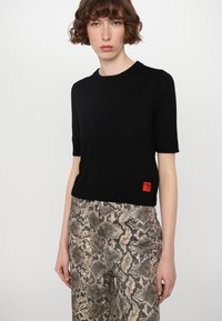 Top corto a maniche corte nero in maglia con scollo a girocollo e un accento di etichetta rossa. Abbinato a pantaloni beige con motivo a serpente.