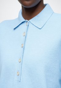 Lichtblauw polohemd met een zachte textuur, voorzien van een kraag en zes lichtgekleurde knopen langs de voorkant.