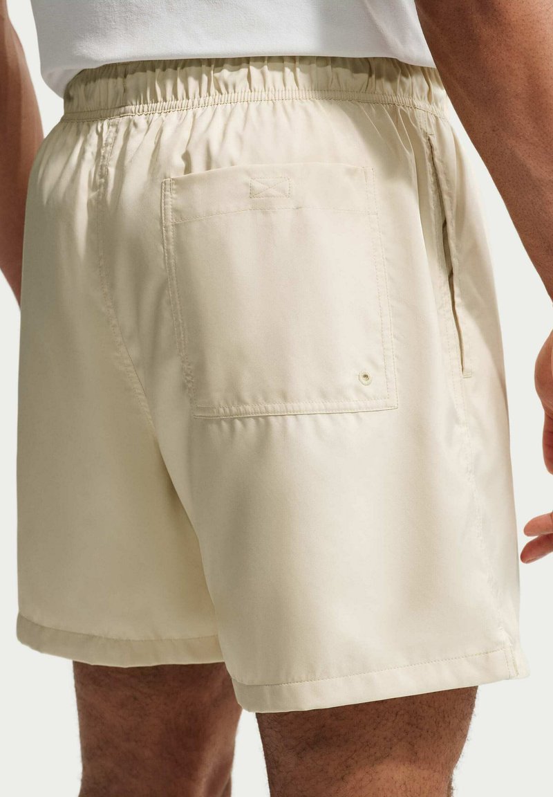 Beige short met elastische taille, achterzak met applicatie en zijnaadzakken, gedragen door een persoon, getoond vanaf halverwege het bovenbeen tot de taille.