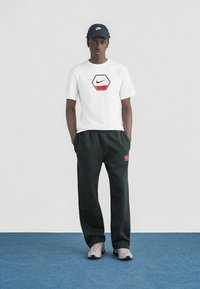 Άνδρας που φοράει λευκό μπλουζάκι Nike, μαύρο παντελόνι φόρμας Nike, γκρι αθλητικά παπούτσια και μαύρο καπέλο Nike, στέκεται σε μπλε χαλί μπροστά σε απλό λευκό φόντο.