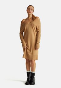Abito lavorato a maglia off-shoulder in un morbido color beige, con texture a righe, maniche lunghe e orlo leggermente svasato. Abbinato a stivali neri con lacci.