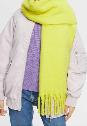 Personne portant une veste bomber rose clair, un pull en tricot violet, un jean bleu et une grande écharpe jaune duveteuse avec des franges pendantes.