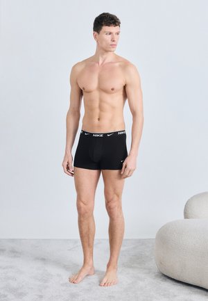 Heas vormis mees seisab paljajalu vaibal, kannab musti Nike bokserlühikuid, vaatab vasakule minimalistlikus toas, kus on ümardatud tool.