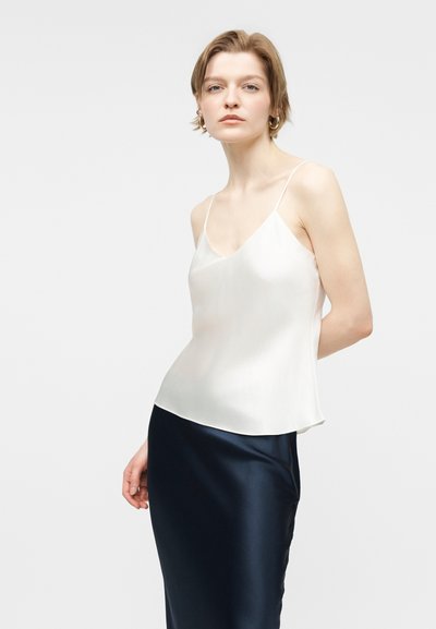 Νεαρή γυναίκα με κοντά μαλλιά, φορώντας λευκό σατέν τοπ με τιράντες και σκούρα ναυτική σατέν φούστα, μπροστά σε απλό φόντο.