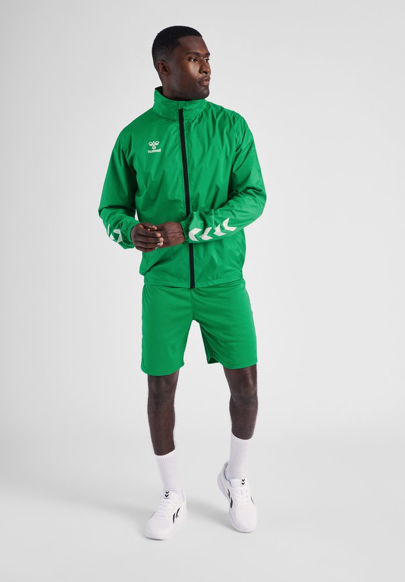 Grön lättviktsjacka med dragkedja, med vit zigzagdesign på ärmarna; matchande gröna shorts; bärs med vita skor och strumpor.