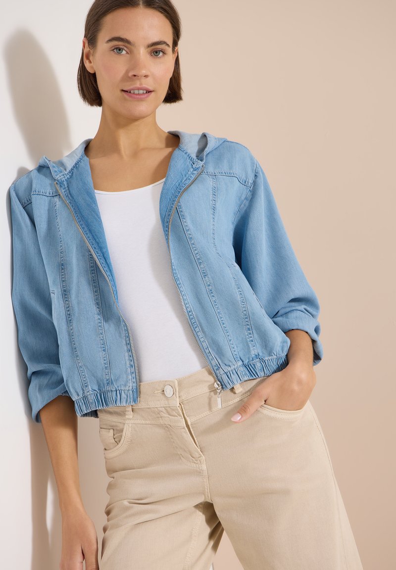 Femme portant une veste en jean courte bleu clair sur un haut blanc, associée à un pantalon beige, debout devant un fond beige.