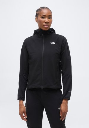 Femme portant une veste à capuche zippée noire North Face et un pantalon noir assorti, sur un fond clair uni.