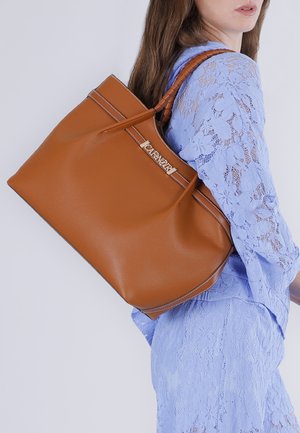 Mujer vestida con un conjunto de encaje azul que lleva un gran bolso de cuero marrón con asa tejida y detalle de logo dorado en el hombro.