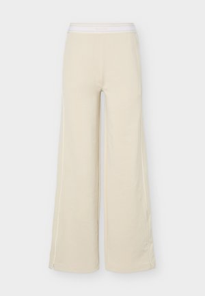 Bež široke hlače s elastičnim pojasom i bijelim obrubom duž vanjskih šavova nogavica, prikazane na jednobojnoj pozadini.