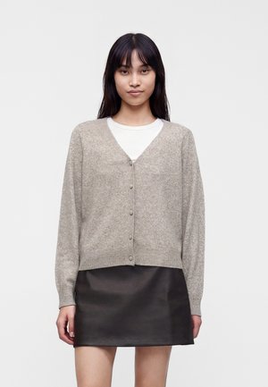 Giovane donna che indossa un cardigan grigio chiaro abbottonato sopra una camicia bianca e una gonna corta nera, in piedi davanti a uno sfondo semplice.