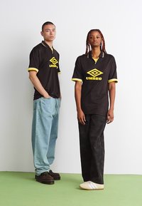 Černé sportovní dresy s žlutými akcenty, se límcem a logem "UMBRO." Modelové mají na sobě světle modré a tmavě vzorované kalhoty.