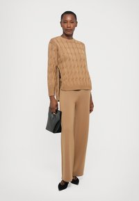 Donna in un maglione beige a trecce e pantaloni a gamba larga che tiene una borsa in pelle nera, indossando tacchi neri, su uno sfondo bianco.